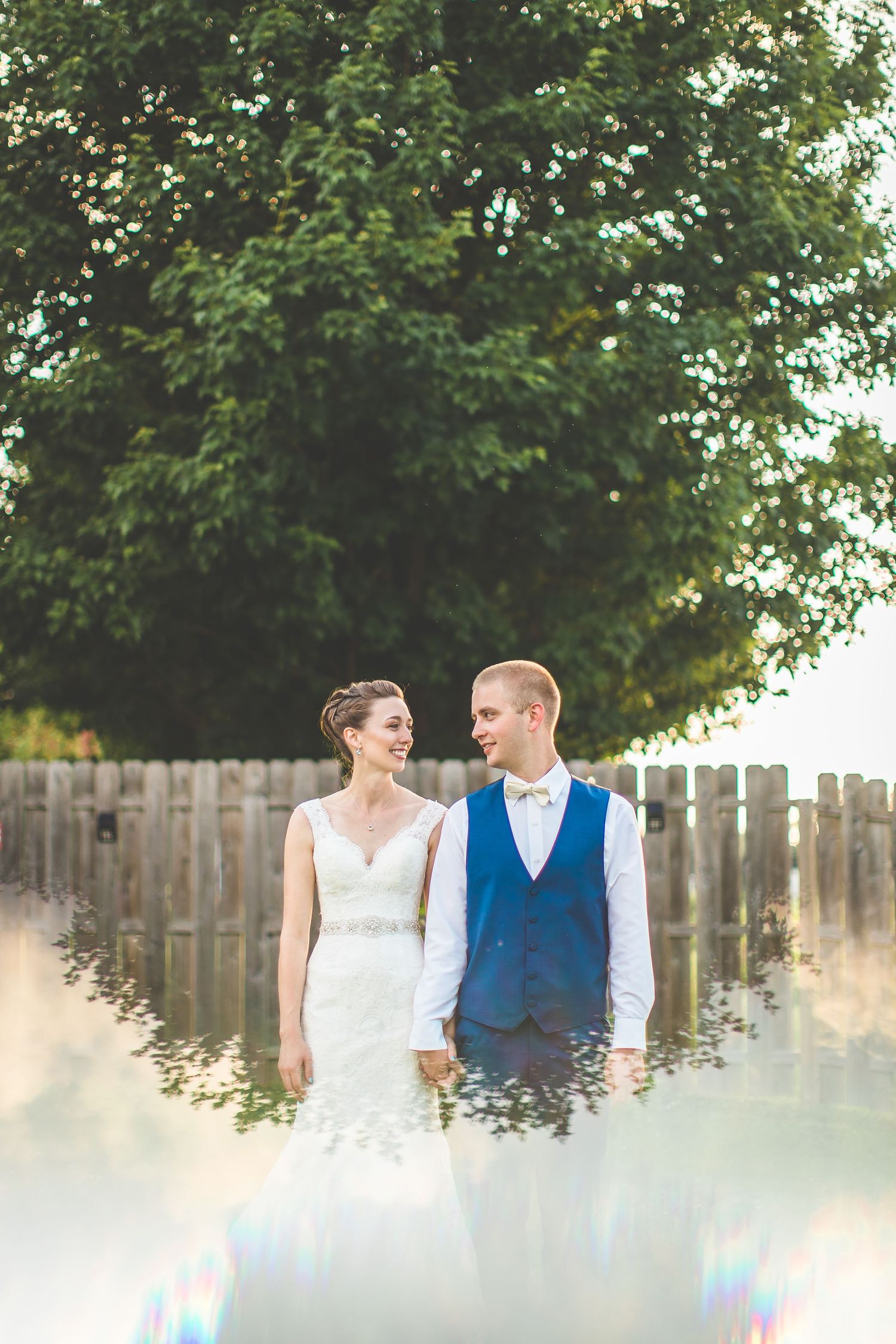 A couple poses together outdoors against a wooden fence in wedding attire.