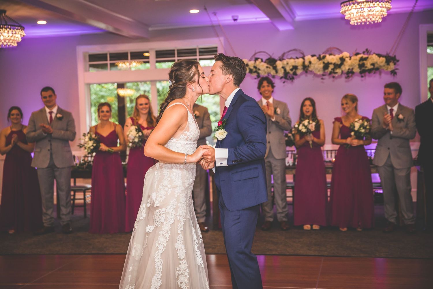 Wedding party and guests watching the couple dance in a purple-lit reception hall with chandeliers.