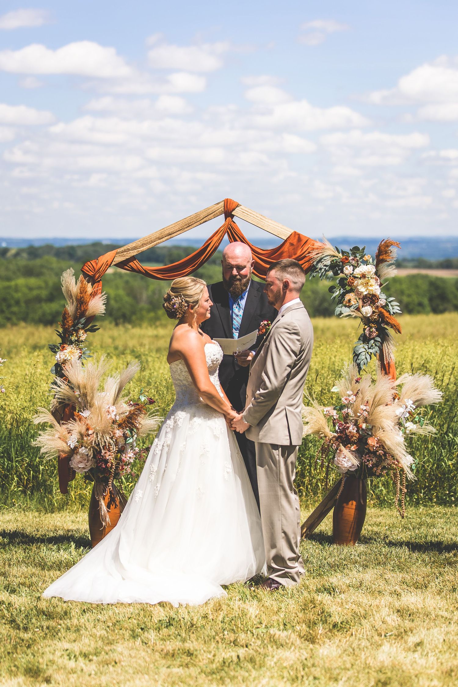 Wedding ceremony under orange fabric arch in scenic hilltop meadow.