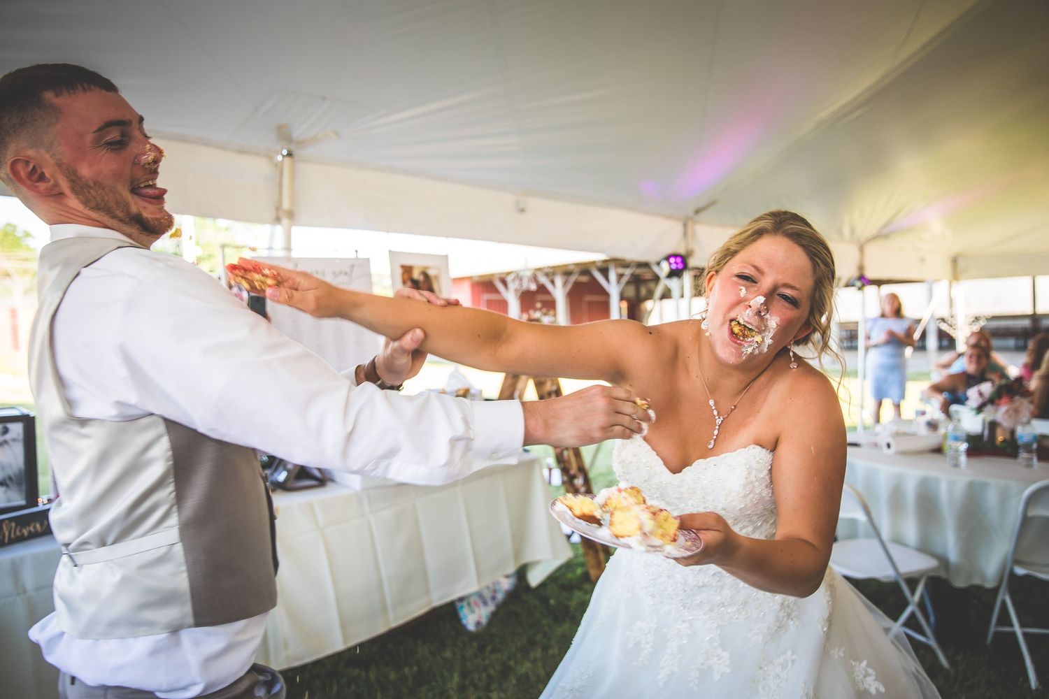 A playful cake-feeding moment between newlyweds under a white tent at their wedding reception.
