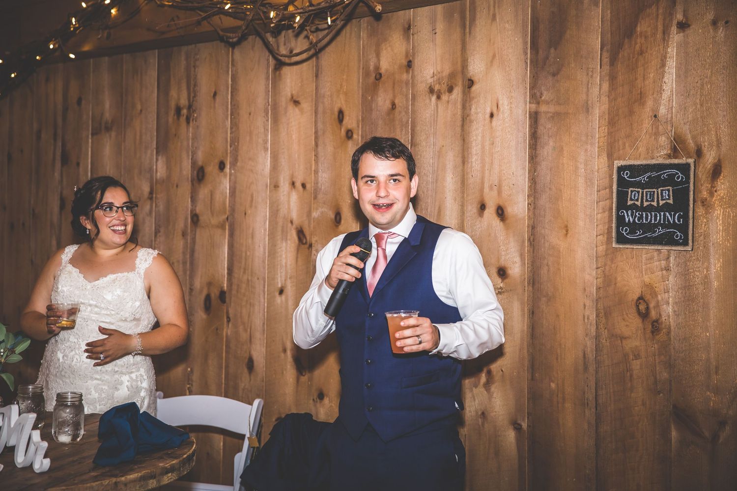 Wedding party enjoys dinner and toasts at rustic barn reception with string lights.