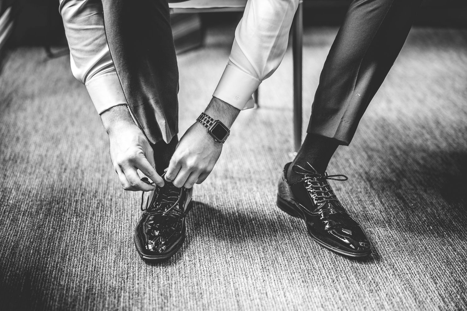 Black and white photo of hands tying formal dress shoes on carpeted floor.