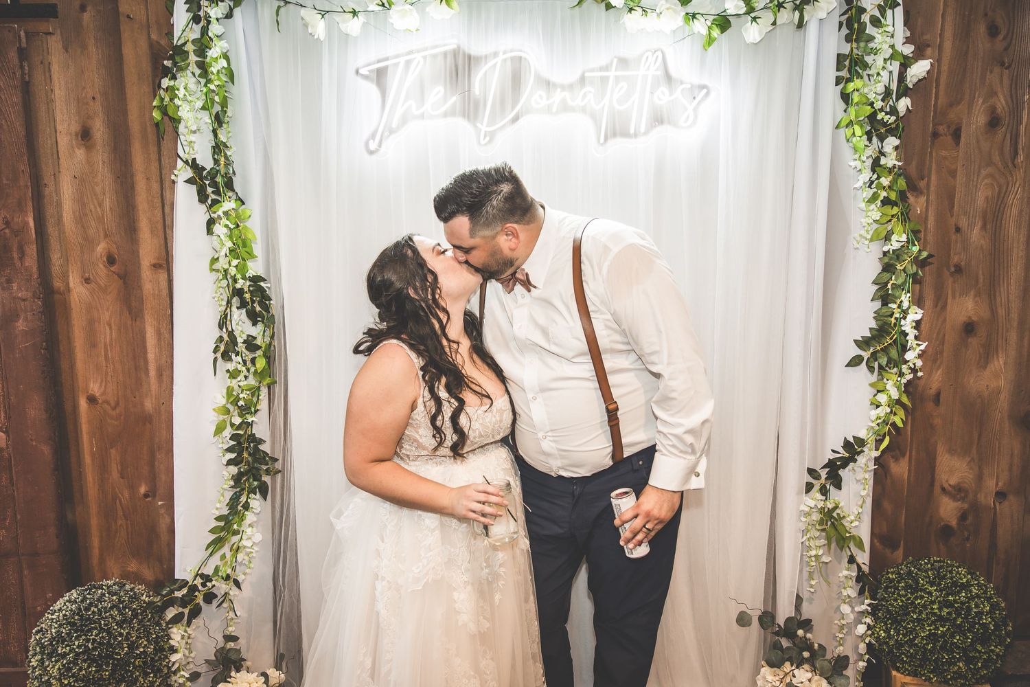 Wedding couple shares romantic moment under greenery arch with white backdrop.