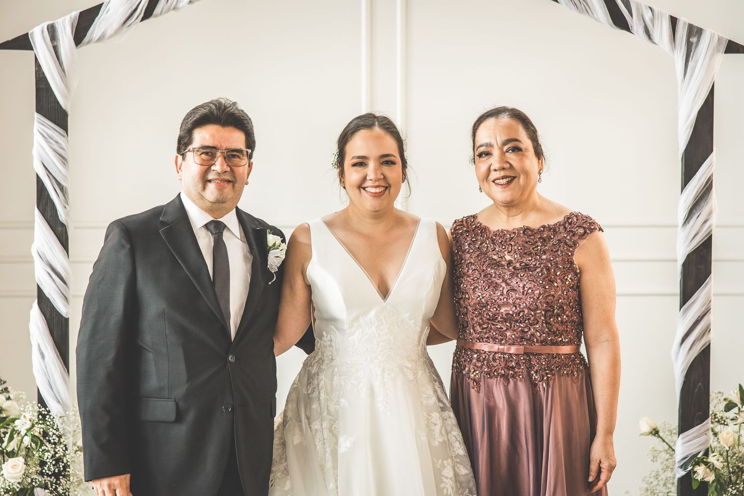 Wedding party poses together in formal attire including burgundy and red dresses against a white wall.