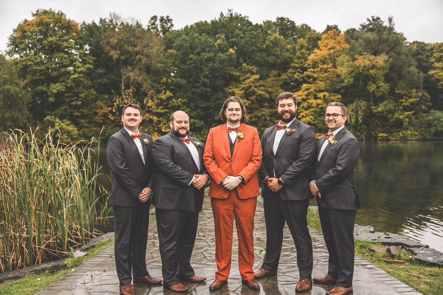 Wedding party poses by a lake with groomsmen in black suits and one in a striking orange suit on an autumn day.