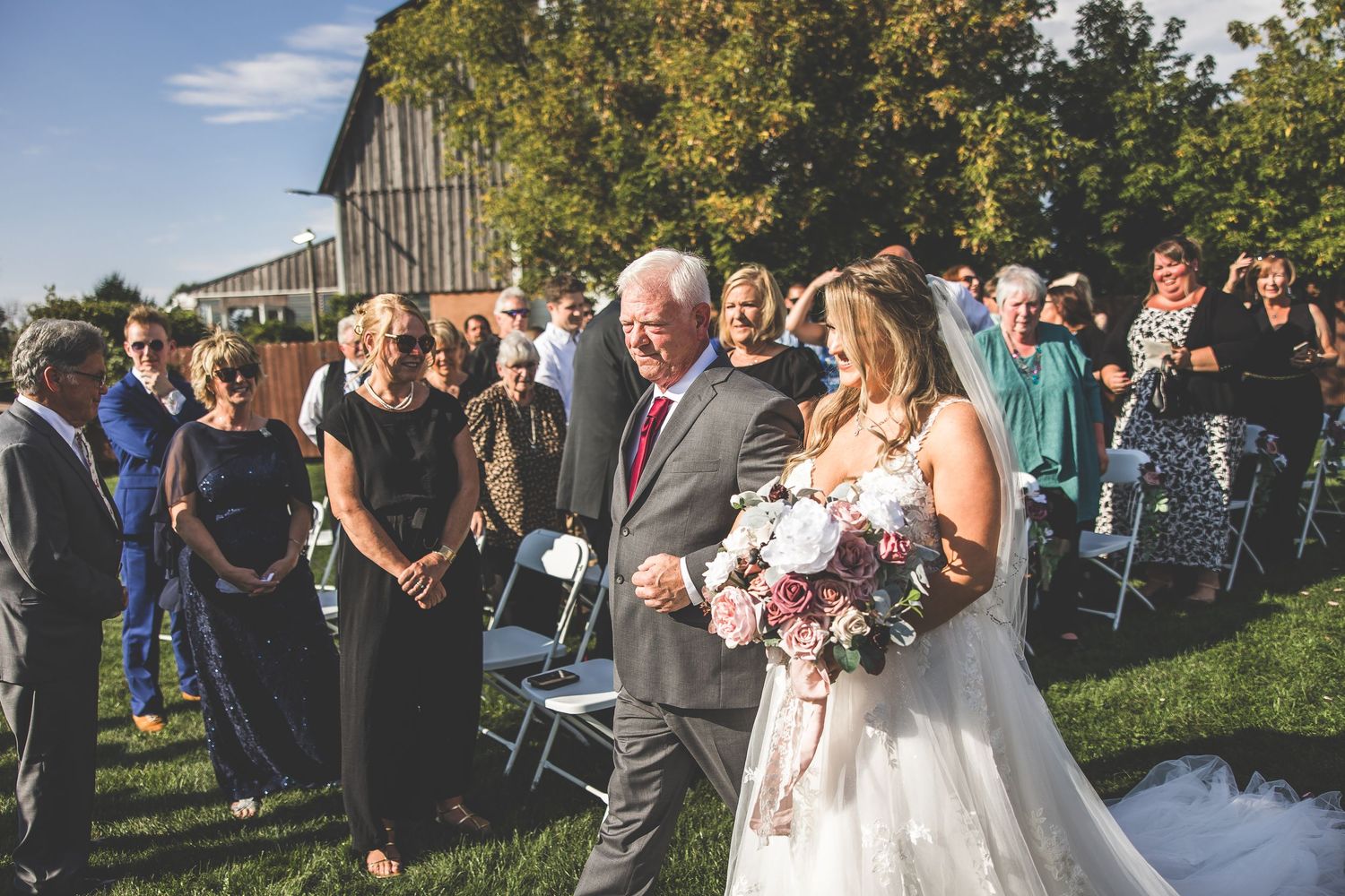 Wedding ceremony processional with guests standing outdoors near rustic barn venue.