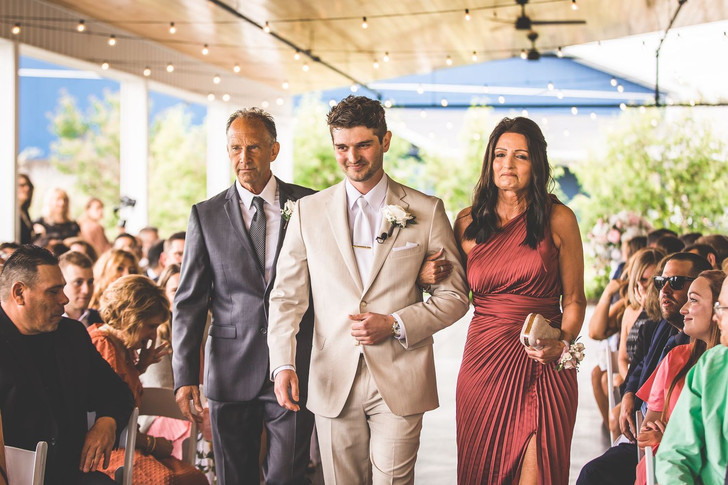Wedding ceremony guests gathered under a covered outdoor venue with string lights.
