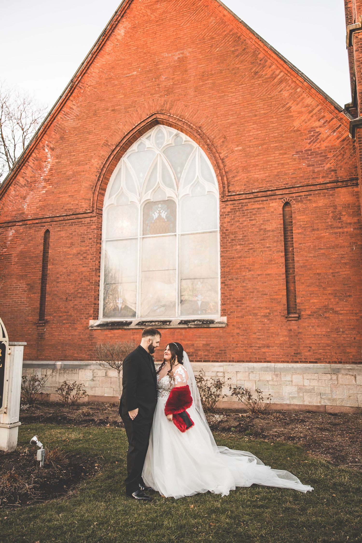 Wedding portraits against red brick church with gothic windows at sunset.