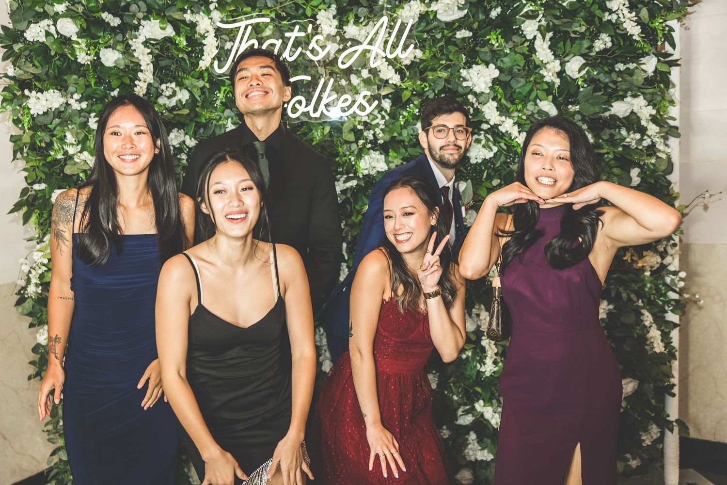 Wedding party poses against a lush floral wall backdrop wearing burgundy and navy attire.