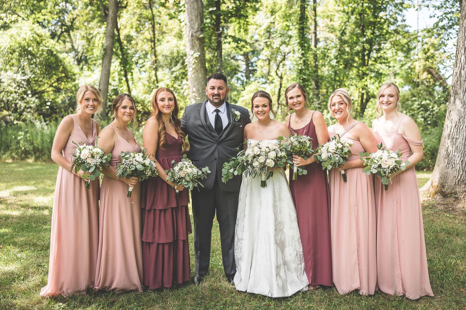 Wedding party in pink and burgundy dresses poses together on a sunny day among trees.