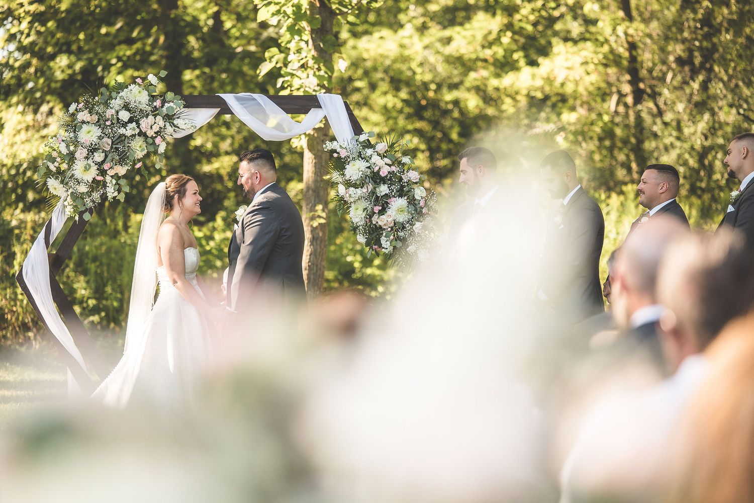 Sunlit wedding ceremony through trees with white draping and floral decorations.