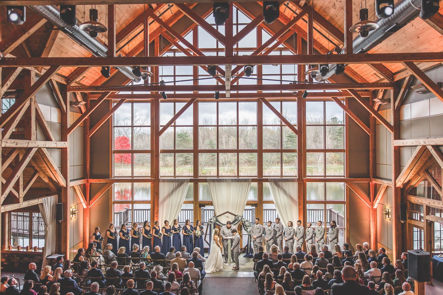 Wide view of wedding ceremony in a light-filled venue with rustic wooden trusses and white draping.