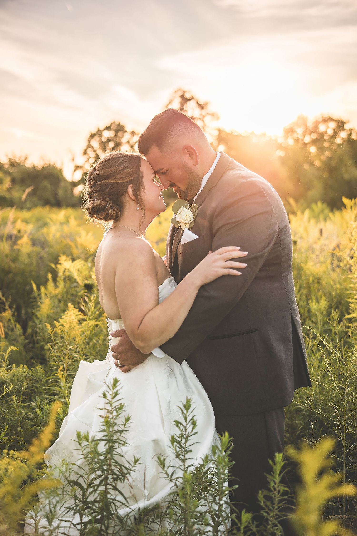 Intimate wedding couple embracing in a meadow during golden hour with sunlit flowers surrounding them.