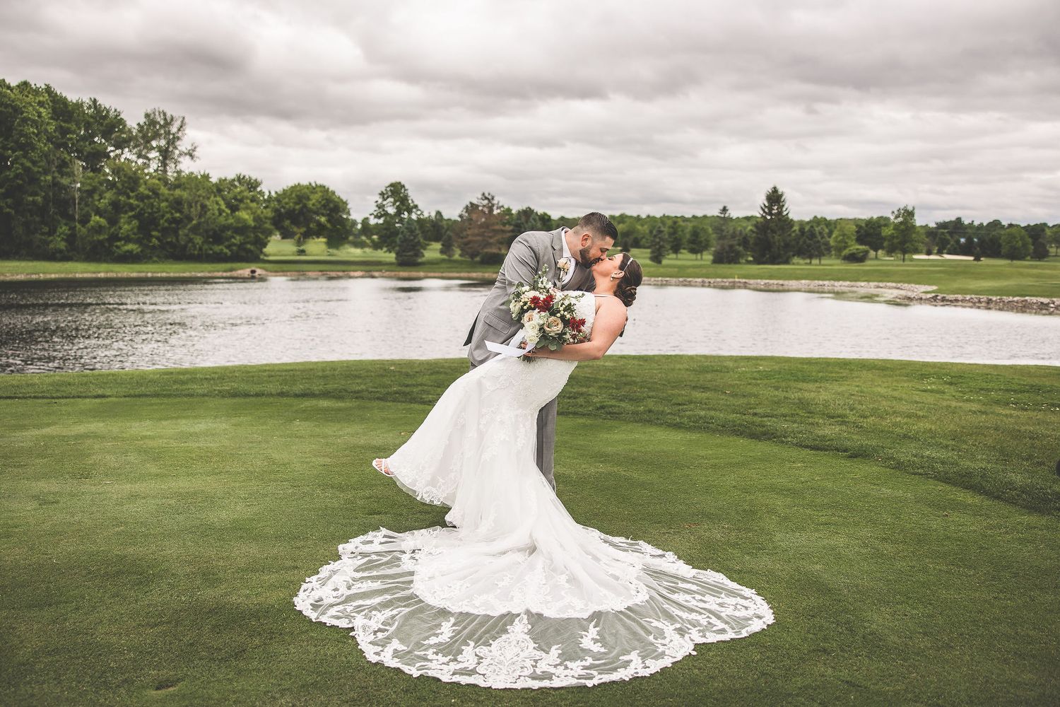 Bride's elegant lace train flows across green grass during lakeside wedding portraits.
