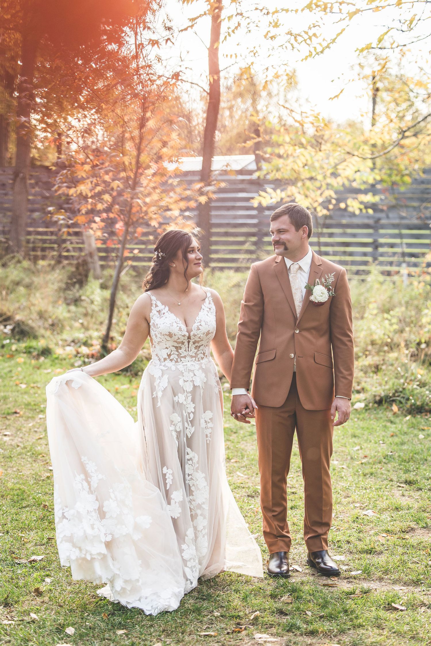 Bride in lace gown and groom in brown suit pose for romantic photos in an outdoor fall setting.