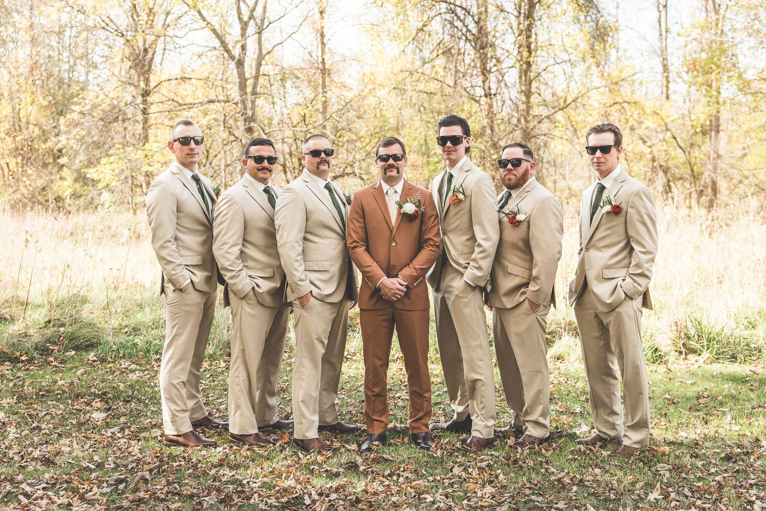 A wedding party of groomsmen in tan suits stands together in a fall woodland setting.