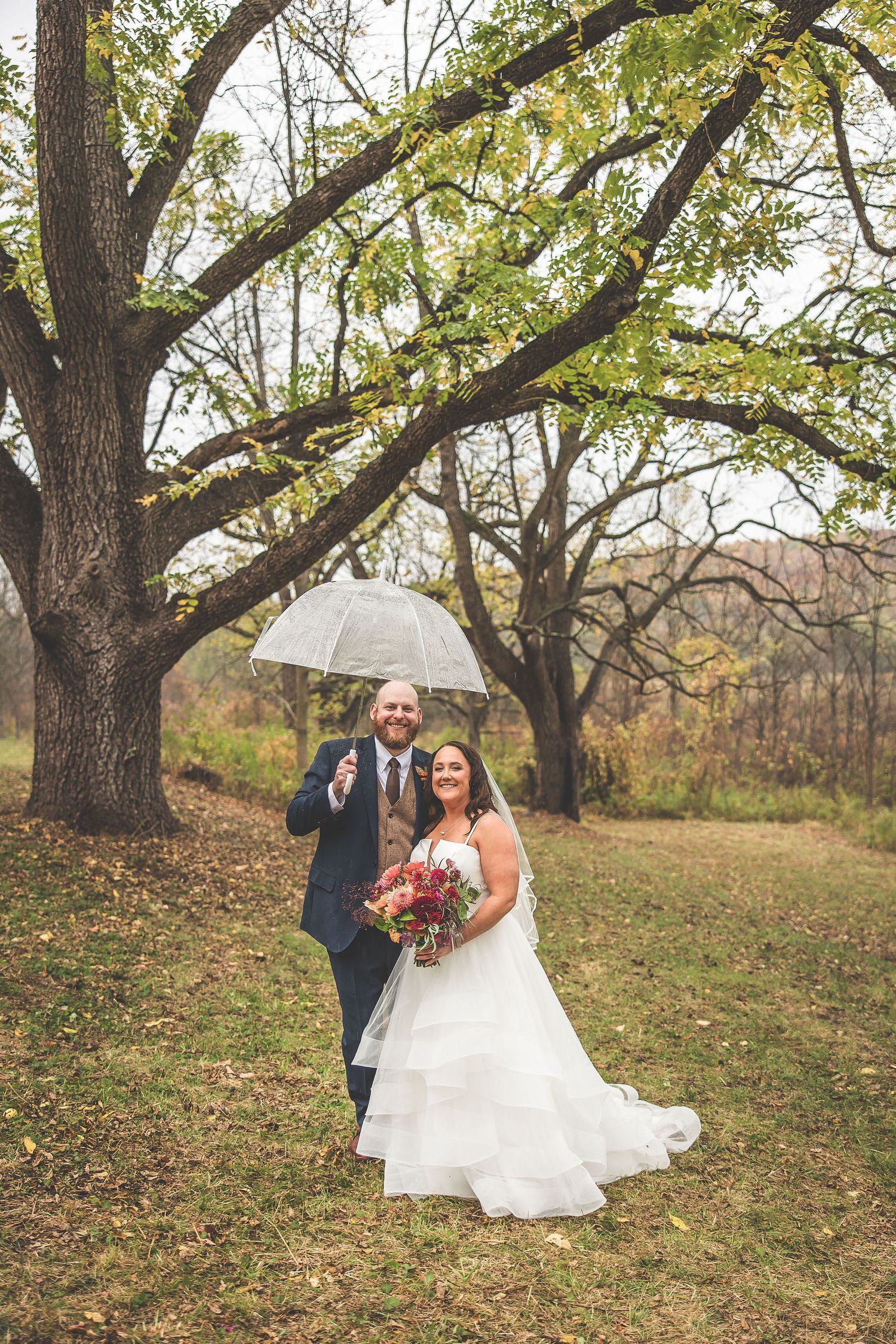 Couple laughs together during outdoor autumn wedding photos beneath tall trees on a hillside.