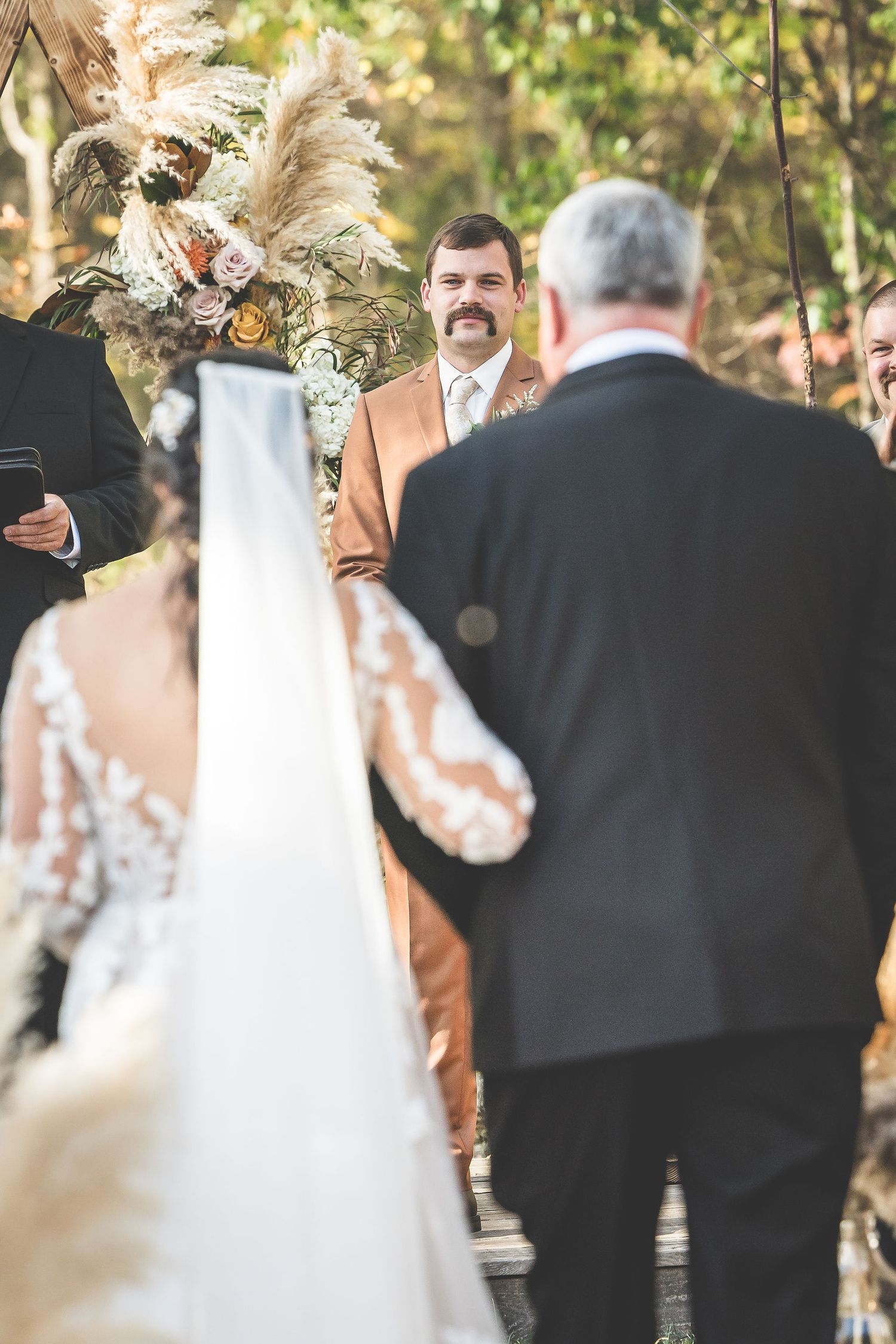 Wedding ceremony attendees gather in a wooded setting with autumn foliage.