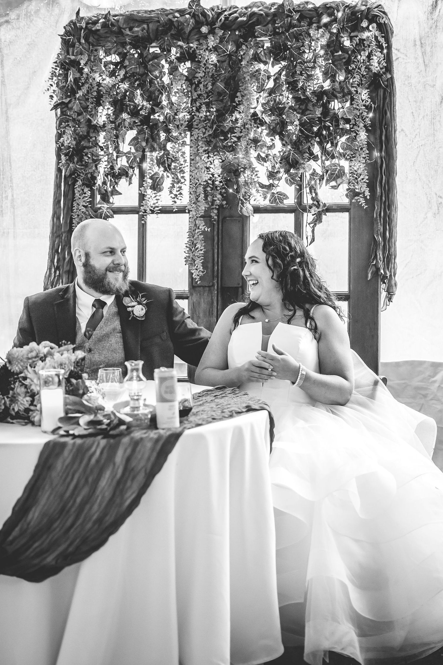Wedding couple seated at decorated head table with floral arrangements and greenery.
