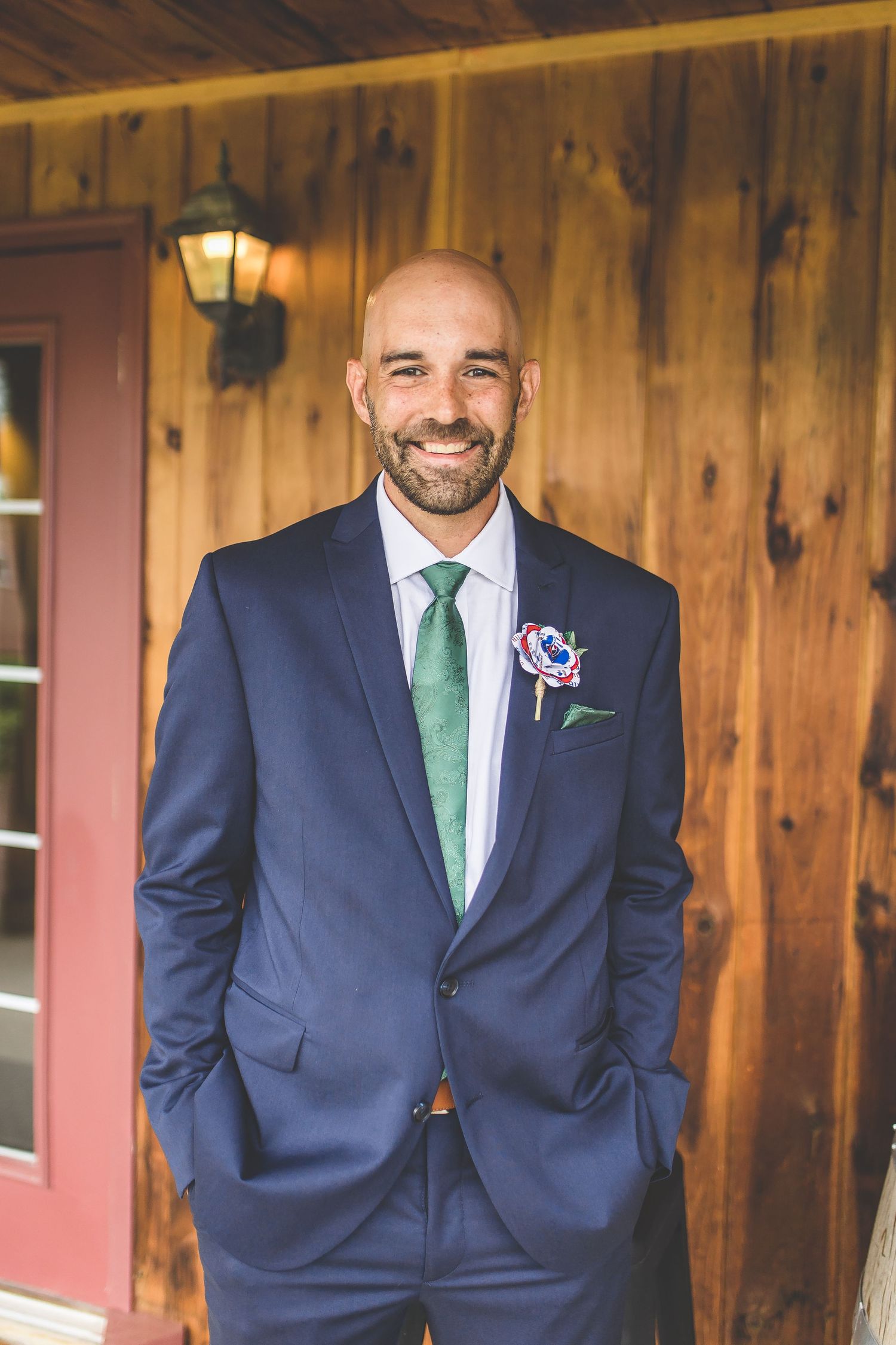 Groomsman in navy suit and green tie poses against rustic wooden wall.