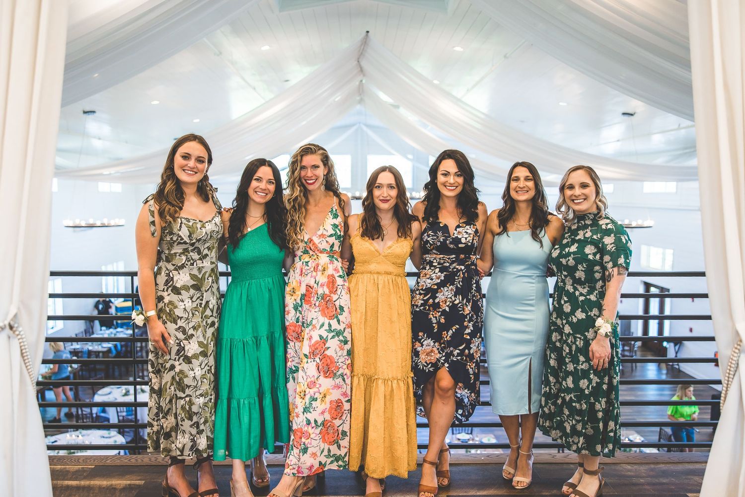 Bridesmaids stand together wearing colorful floral and solid dresses in a white draped venue.