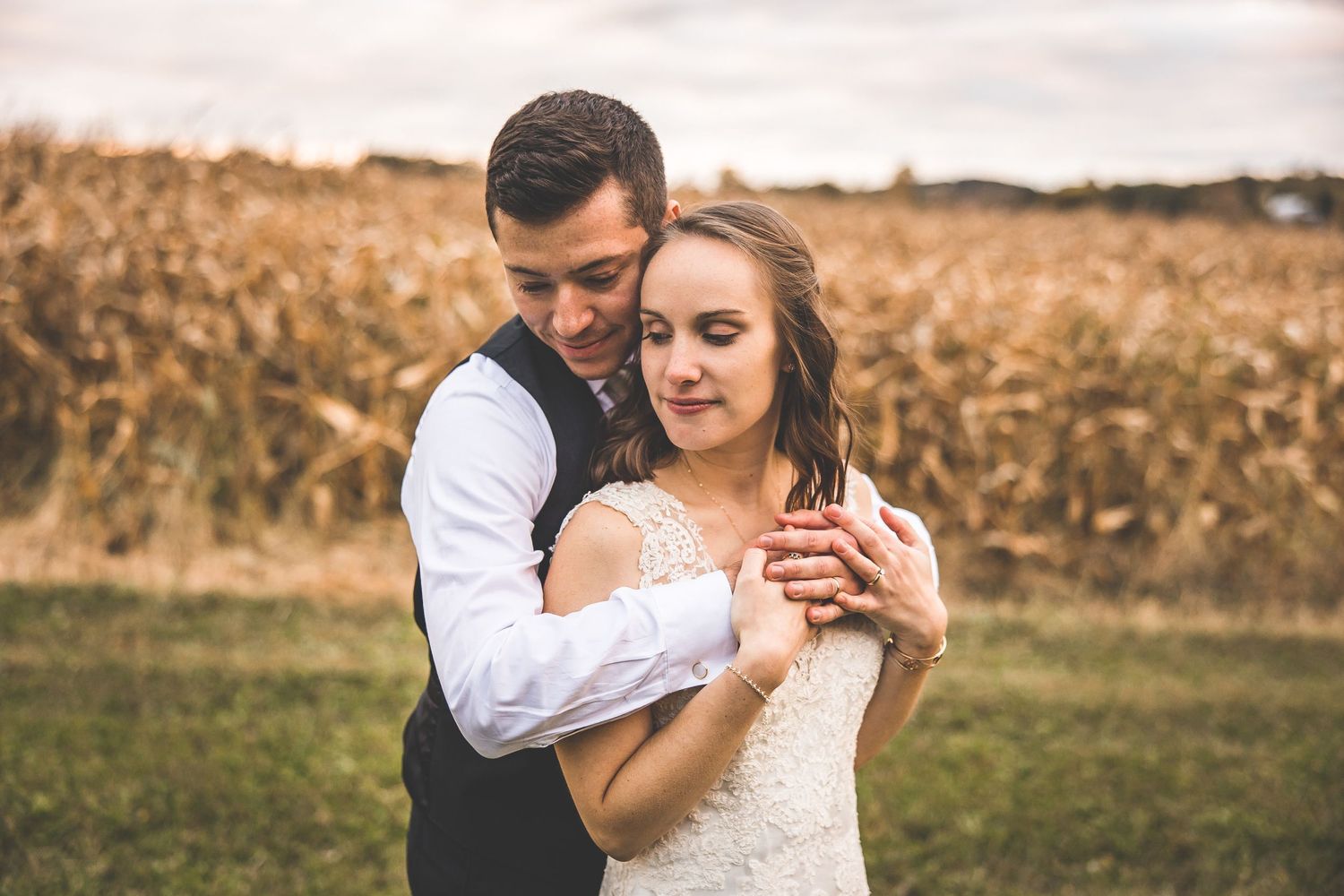 A romantic wedding portrait in a cornfield with the couple embracing in formal attire at sunset.