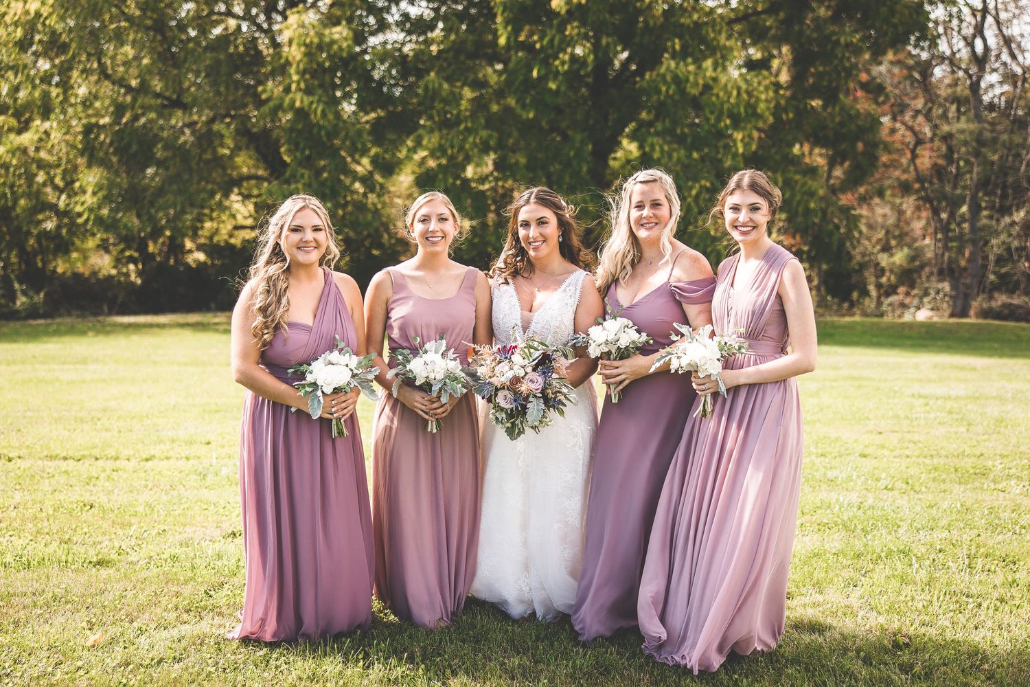Bridal party in dusty rose and mauve dresses pose together on a sunny lawn holding floral bouquets.