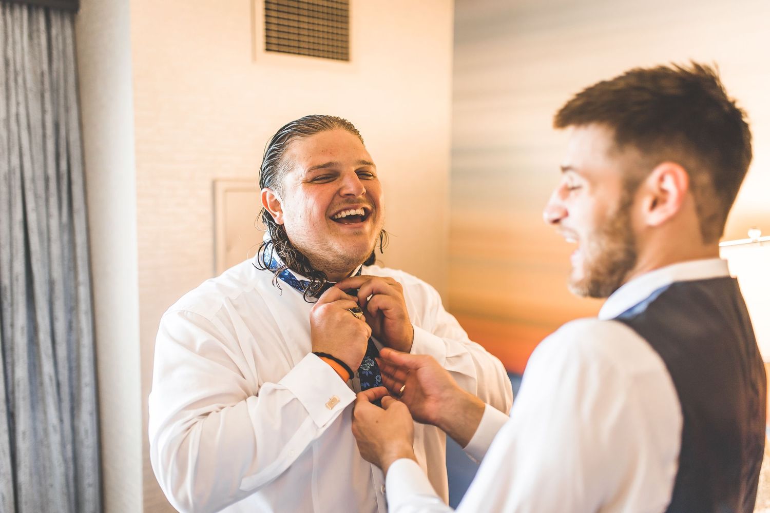 Two people helping with formal attire preparations in a hotel room.