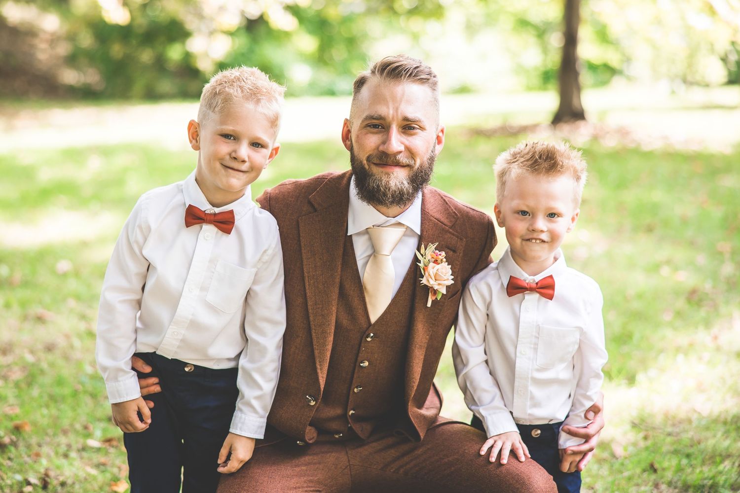 Two young boys in bow ties pose with adult in brown suit at outdoor wedding.