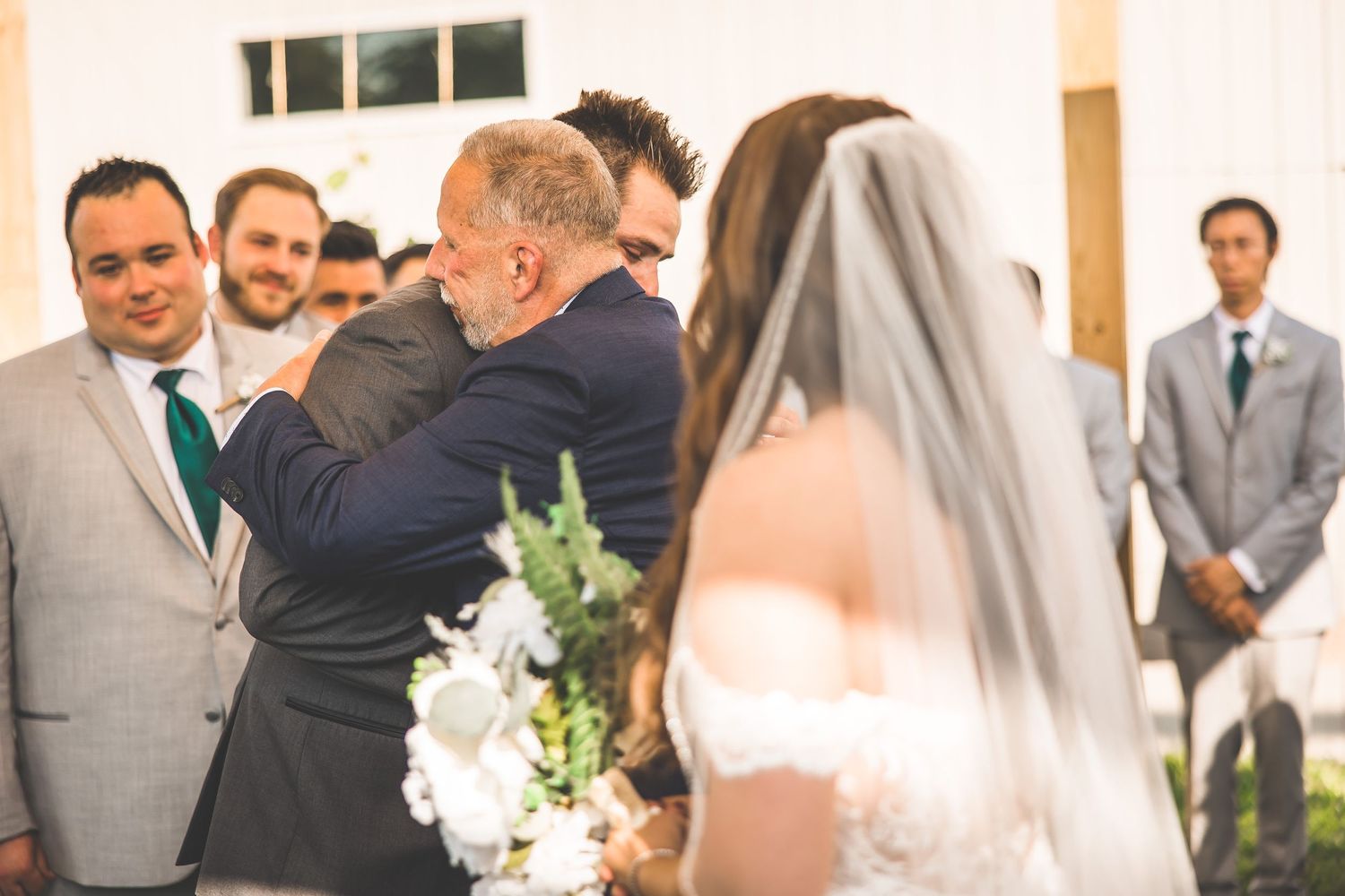 Wedding ceremony participants gathered under a wooden pavilion decorated with white fabric.