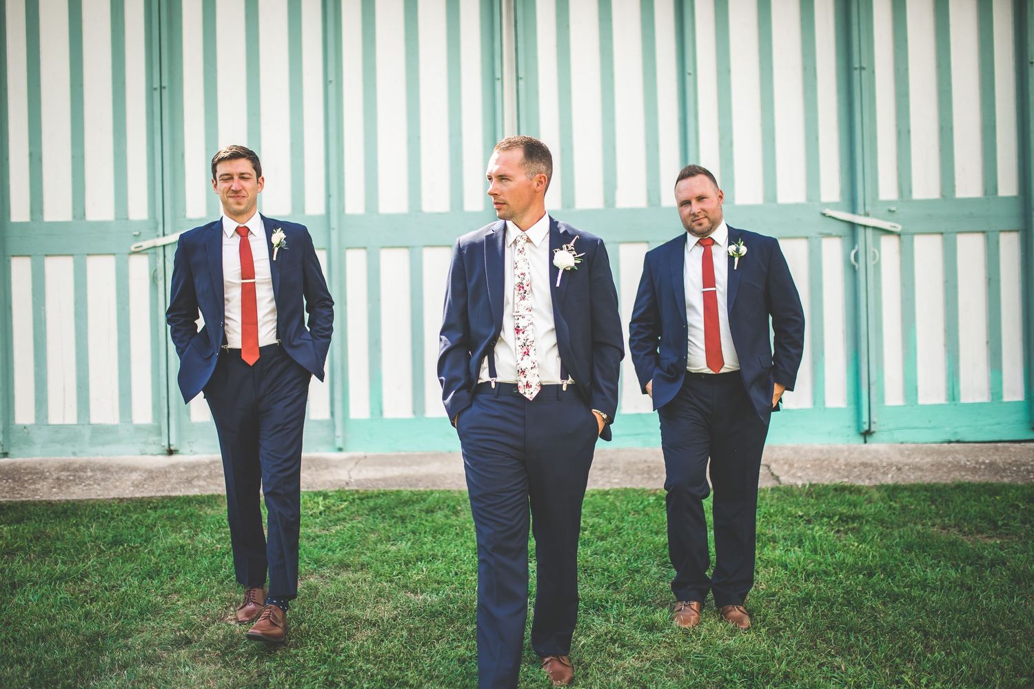 Row of groomsmen in navy suits stand against mint green and white striped building exterior.