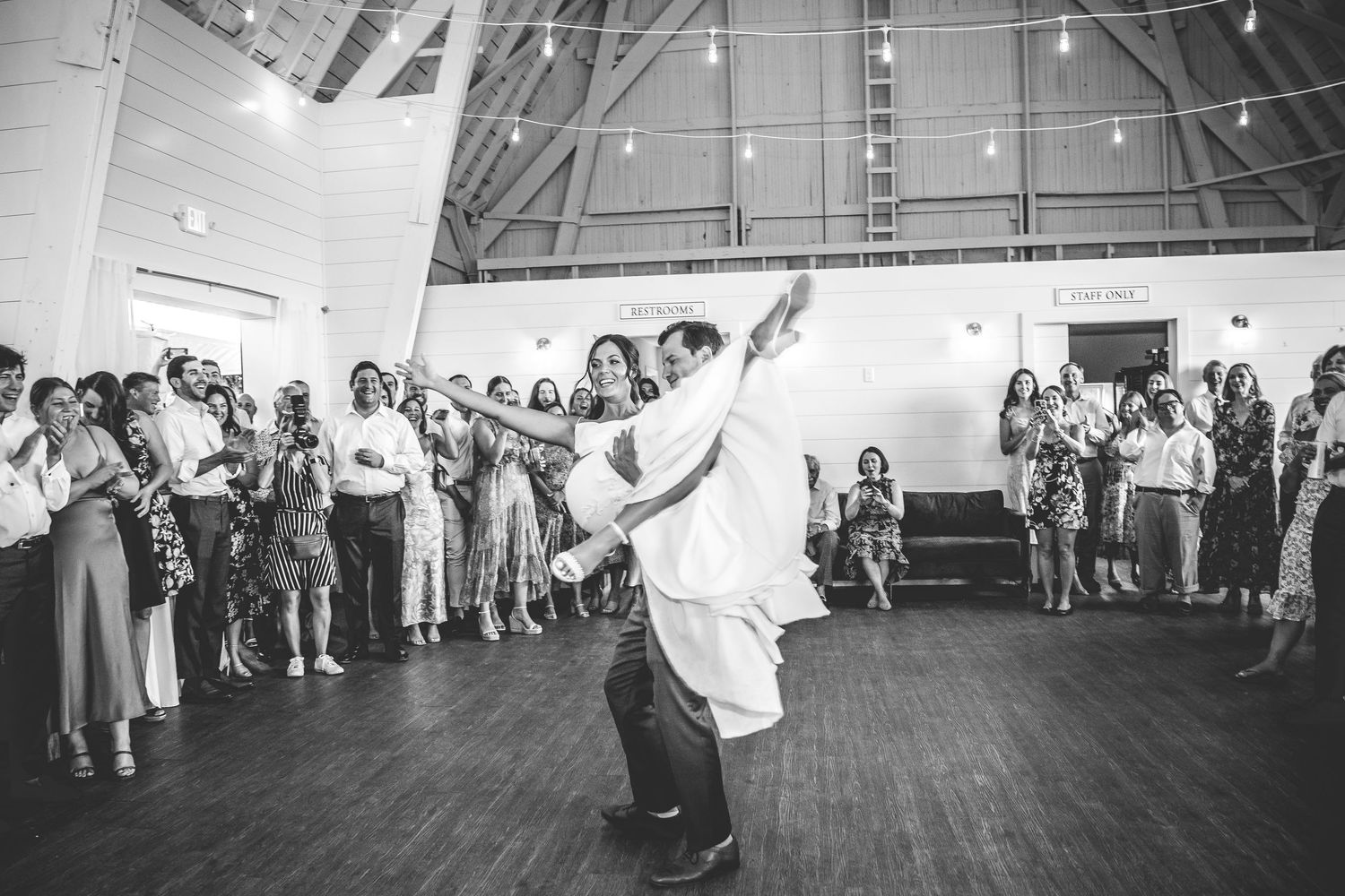 Wedding guests watch as a couple shares their first dance in a rustic venue with wooden floors and white walls.