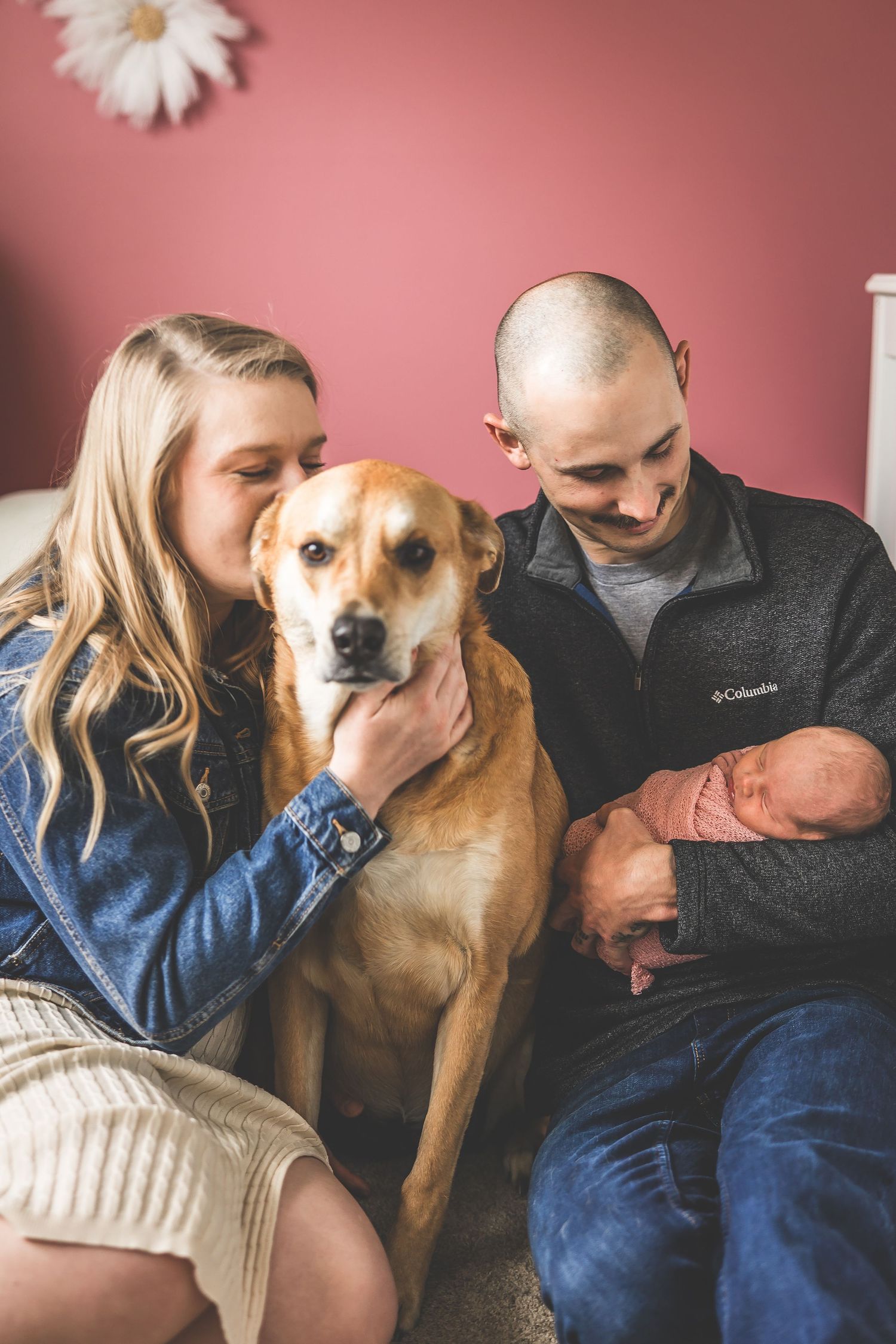 A family cuddles with their tan dog on a cozy couch against a pink wall.