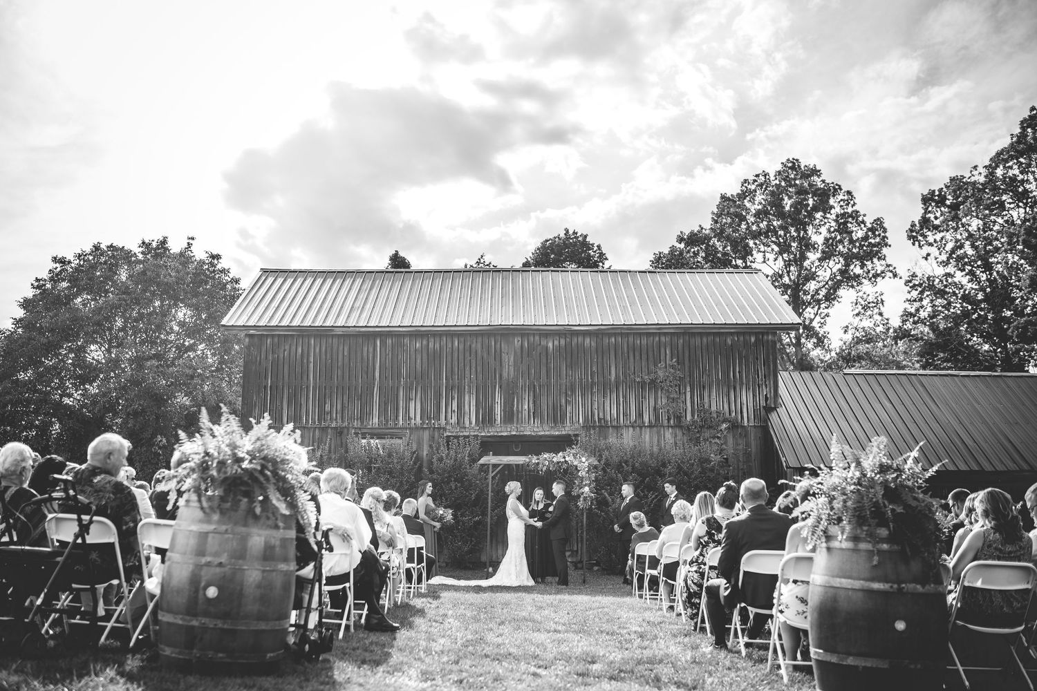 Black and white panoramic view of a rustic barn wedding venue with guests seated outdoors.