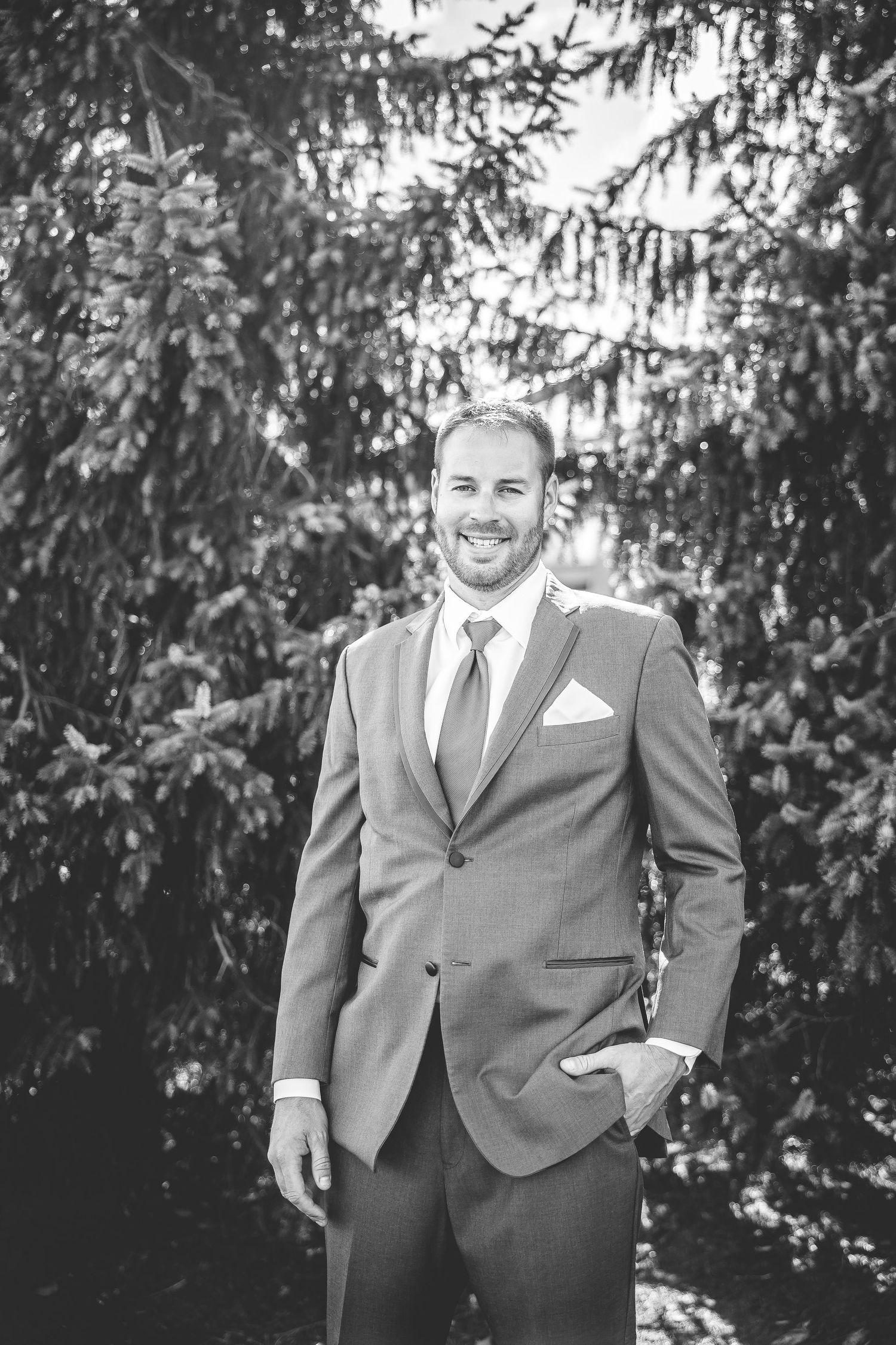Wedding party members in gray suits pose outdoors against evergreen trees.