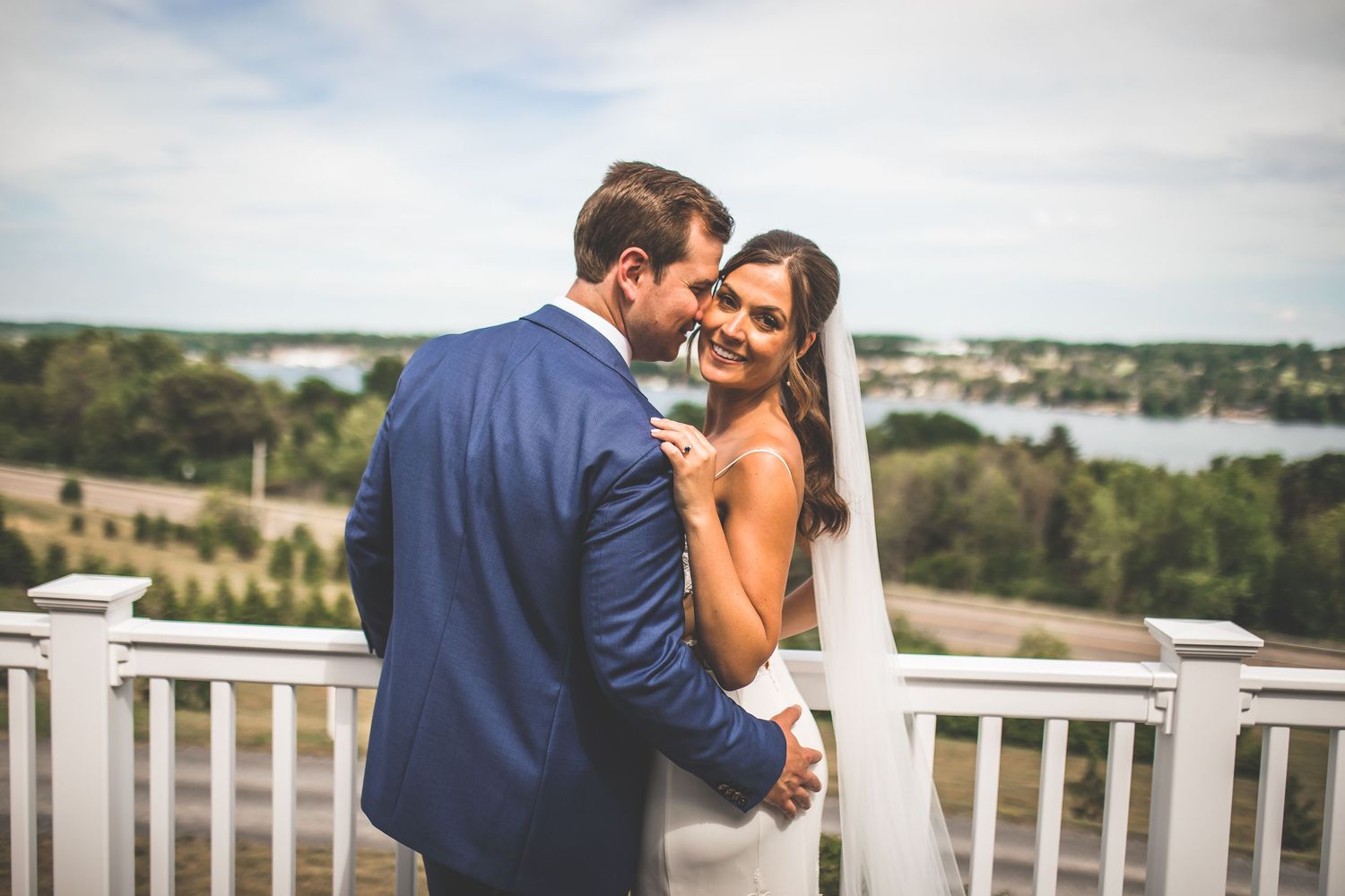 Newlyweds embrace on outdoor balcony with scenic water views in background.