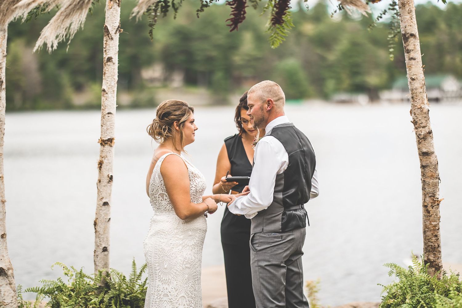 Intimate wedding ceremony moment under a decorated birch arch with lake backdrop and natural lighting.