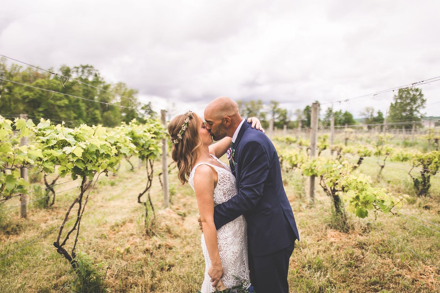 Wedding couple shares tender moments between rows of grapevines on cloudy afternoon.