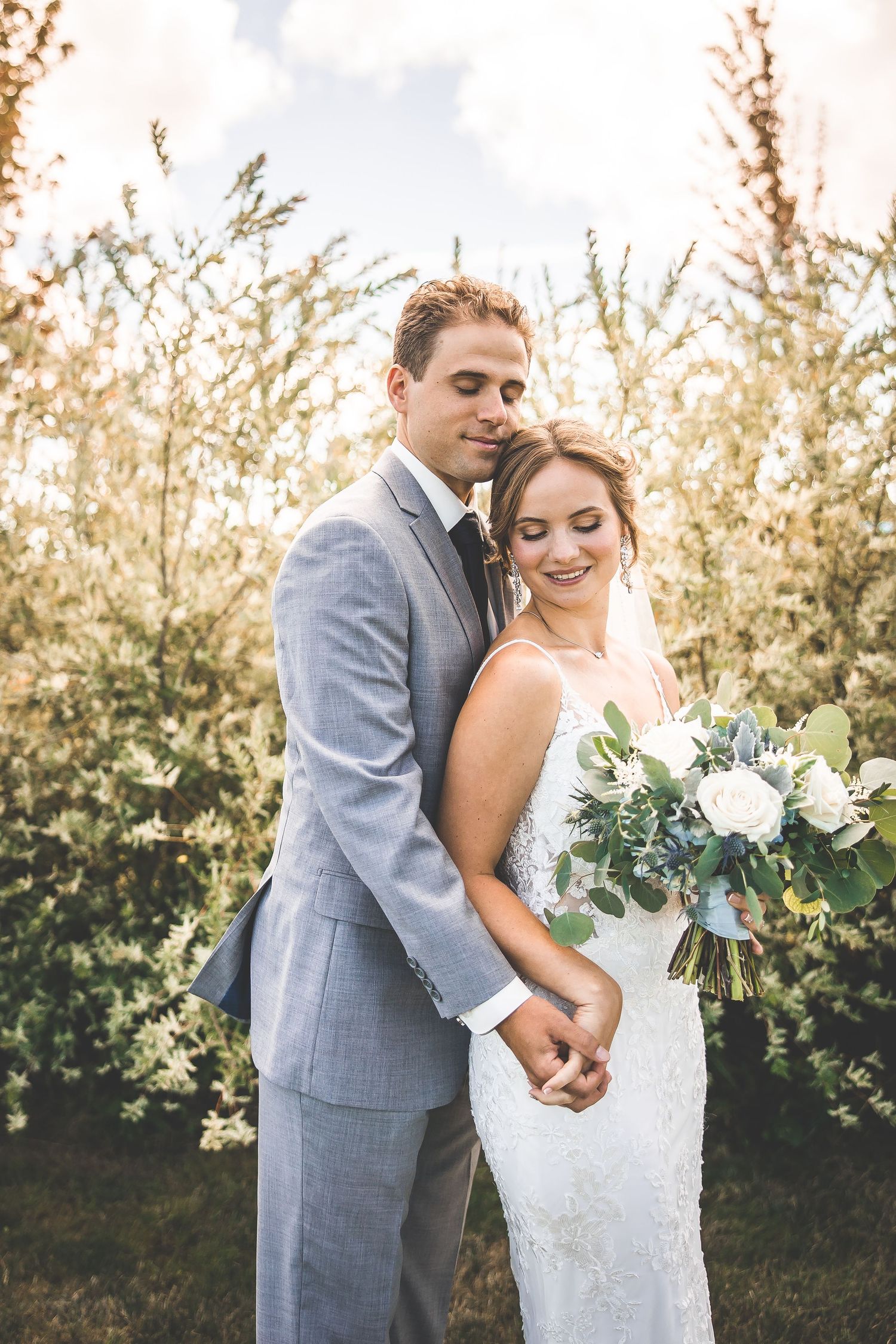 Couple embraces while holding white floral bouquet in front of blooming trees.