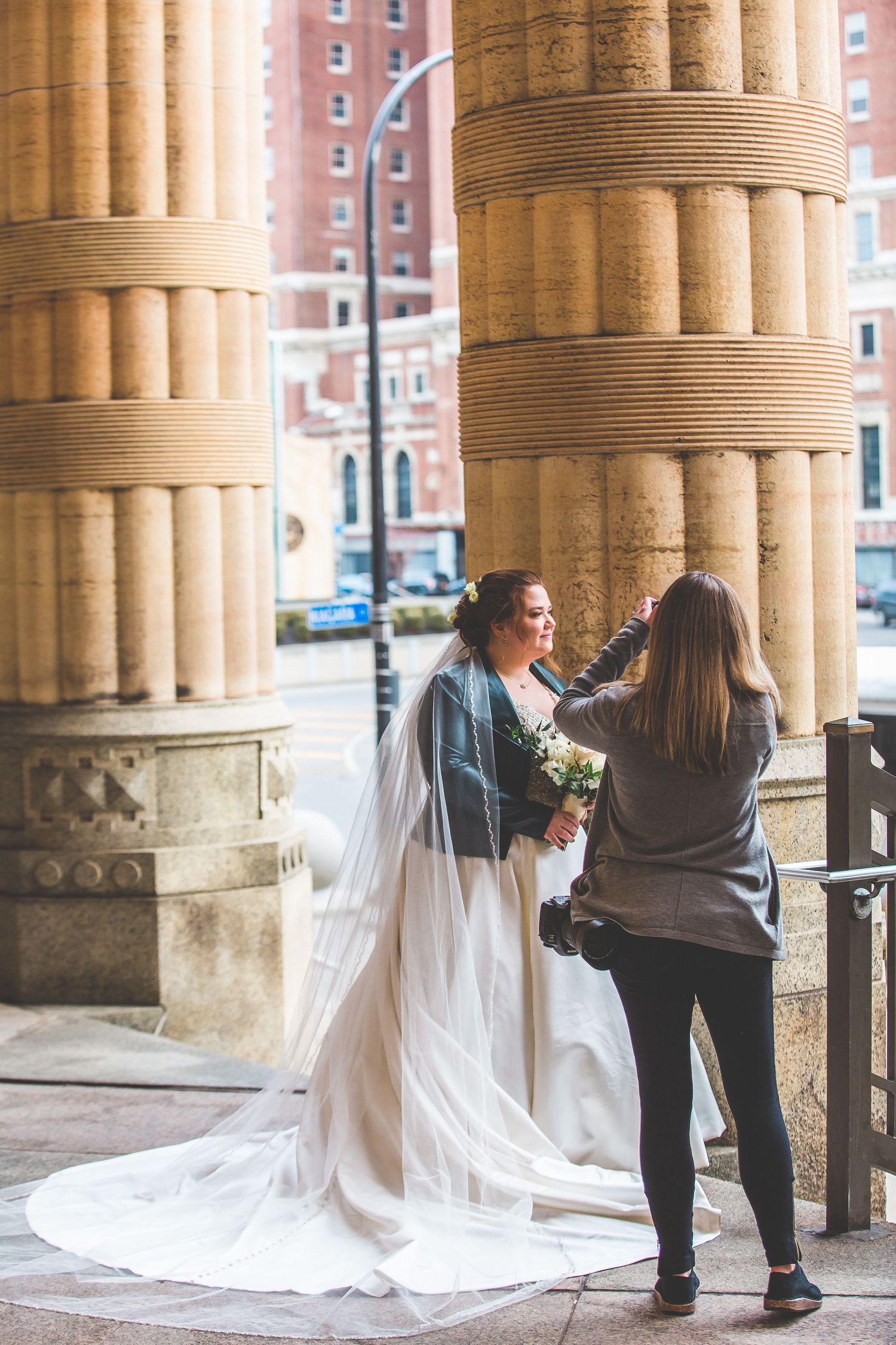 Wedding party arranges bride's flowing white train against classical stone columns.