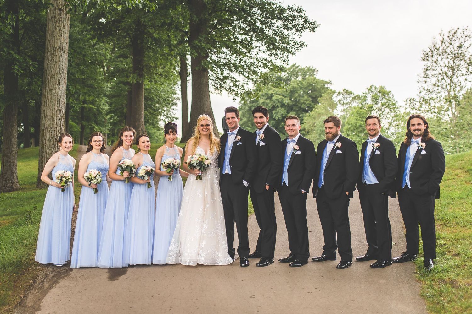 Wedding party in light blue dresses and navy suits poses together on a wooded path.