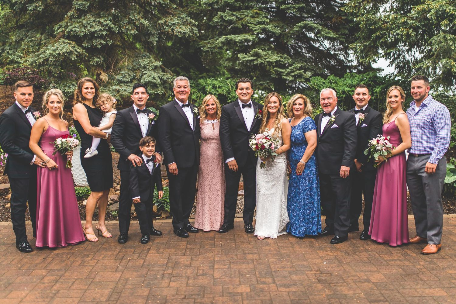 Large wedding party in formal attire posed together on brick patio with evergreen trees in background.