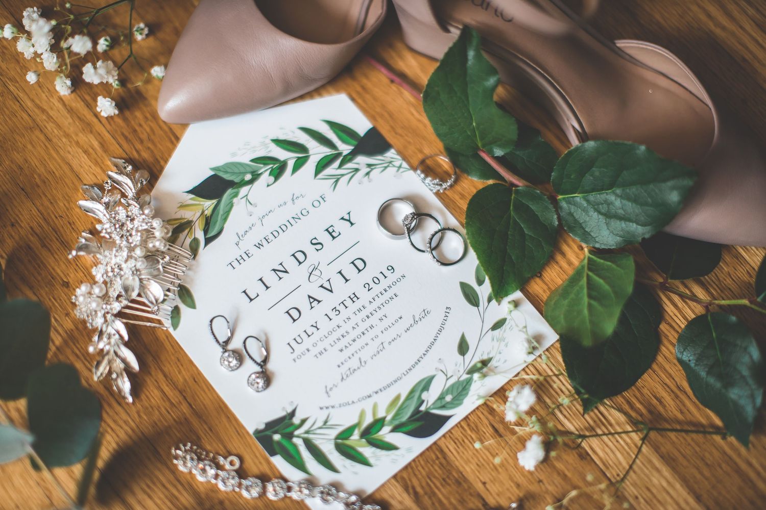 Wedding invitations with green leaf designs and wedding rings arranged on wooden surface.