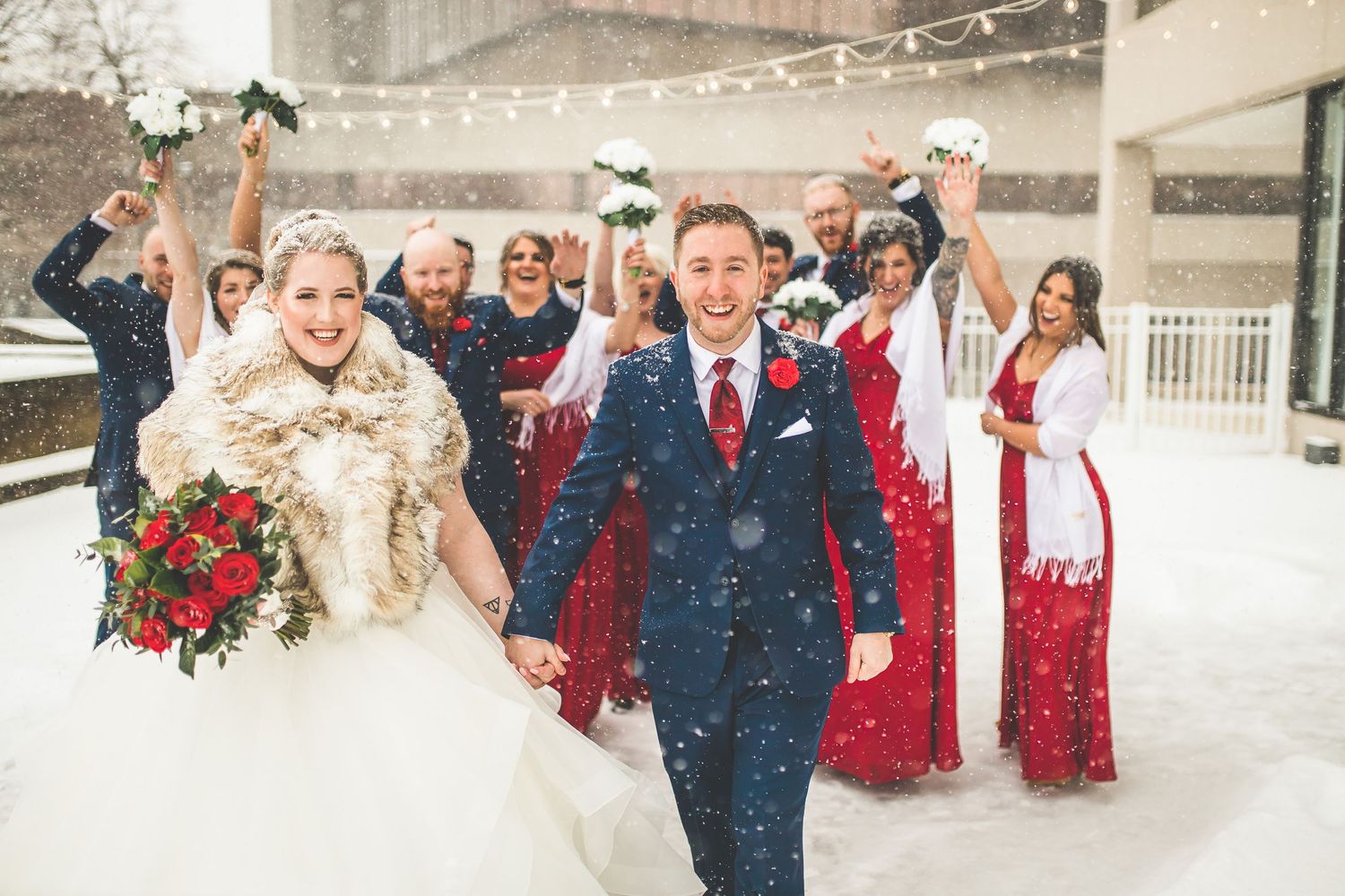 Wedding party celebrates joyfully in snowfall with bridesmaids in red dresses cheering behind couple.
