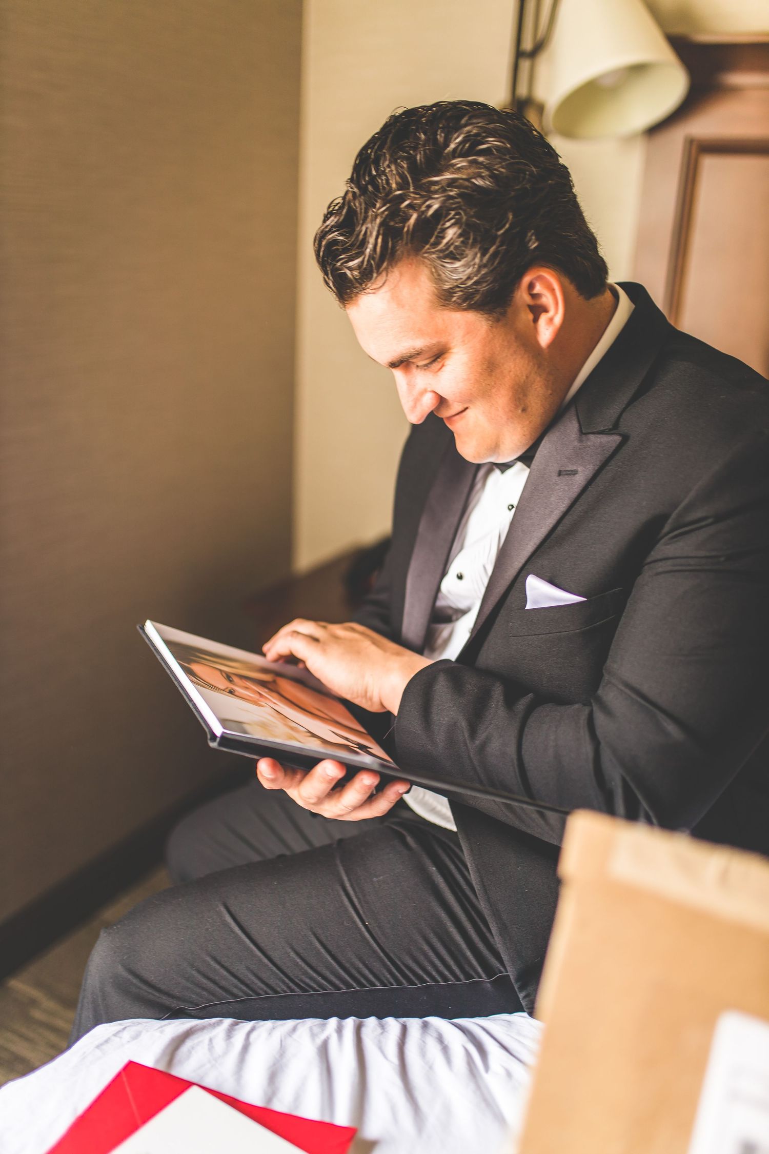 Groom in black tuxedo looking at mobile phone while sitting on bed.