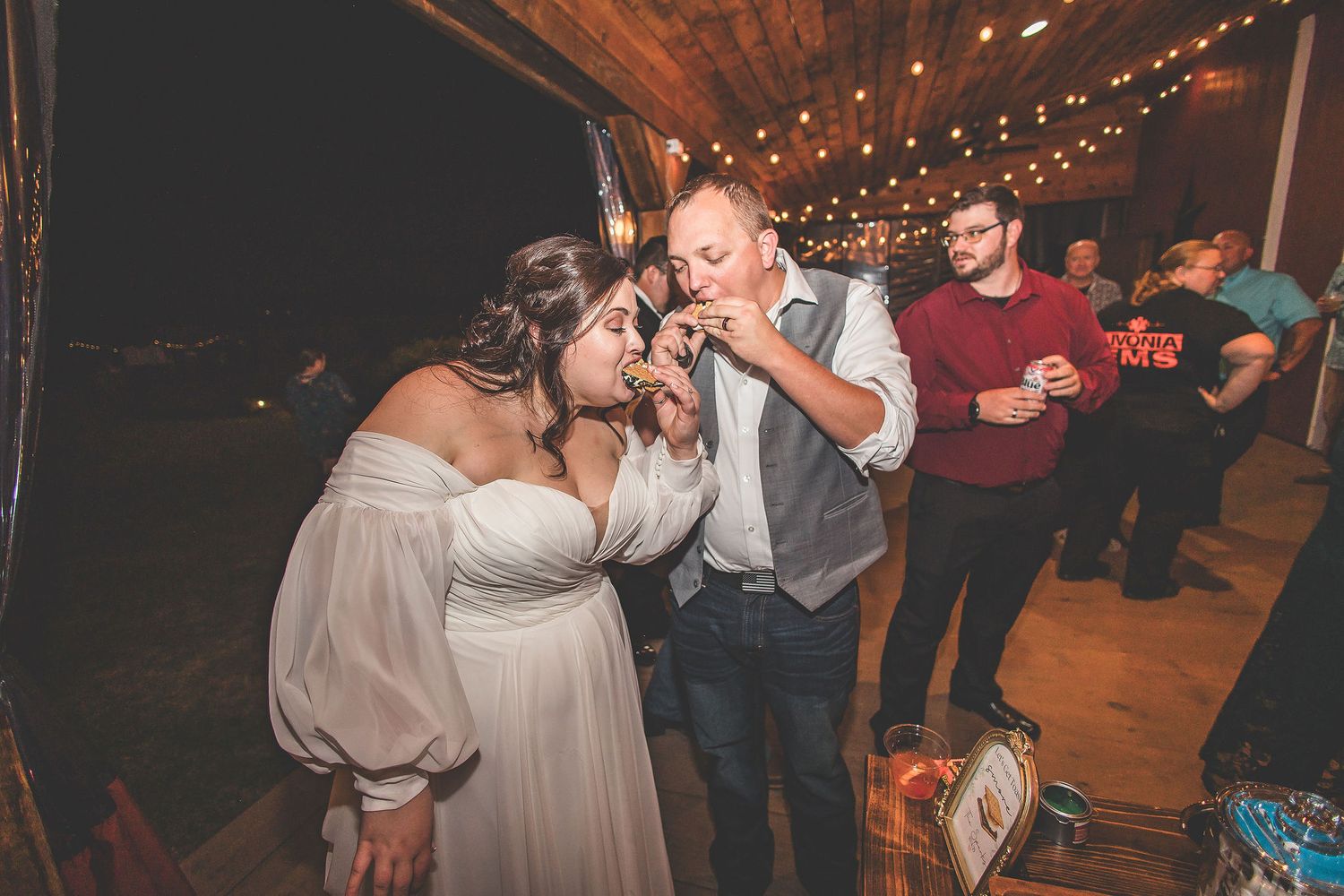 Reception guests gather around a dessert table in a rustic barn setting with string lights.