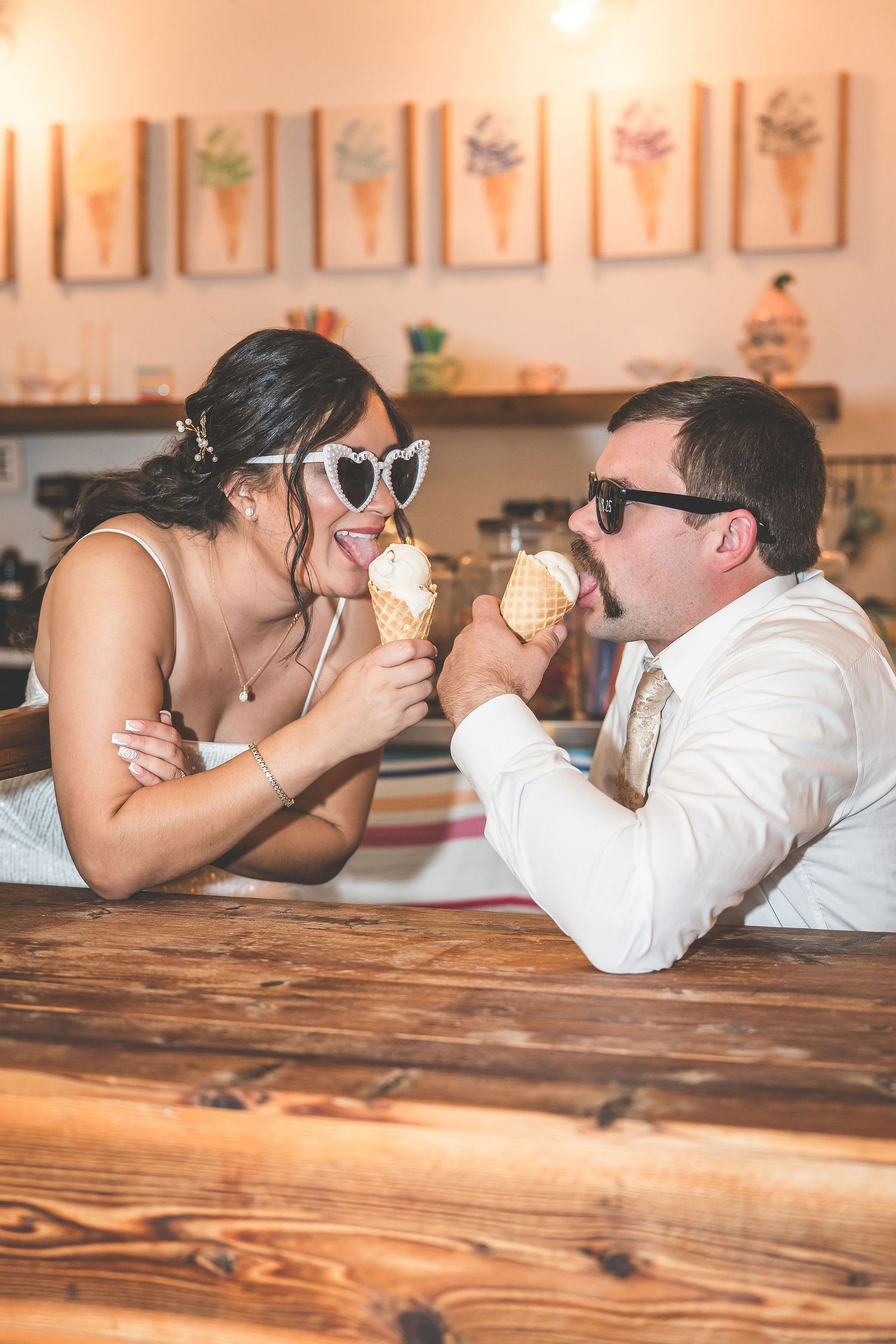 Couple lying on wooden counter eating ice cream cones together in a retro-styled shop.