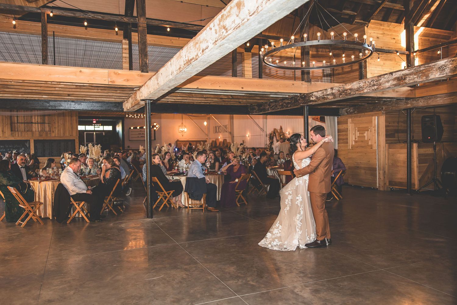 Wedding reception venue with string lights and rustic wooden beams creates warm ambiance for dancing guests.