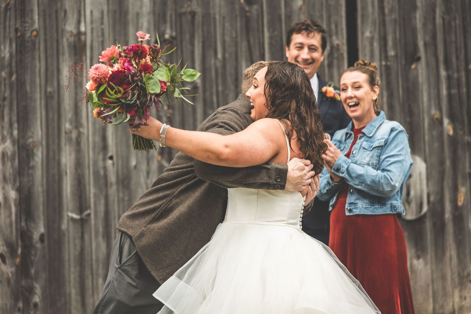 Wedding party celebrates with laughter and joy against wooden barn backdrop.