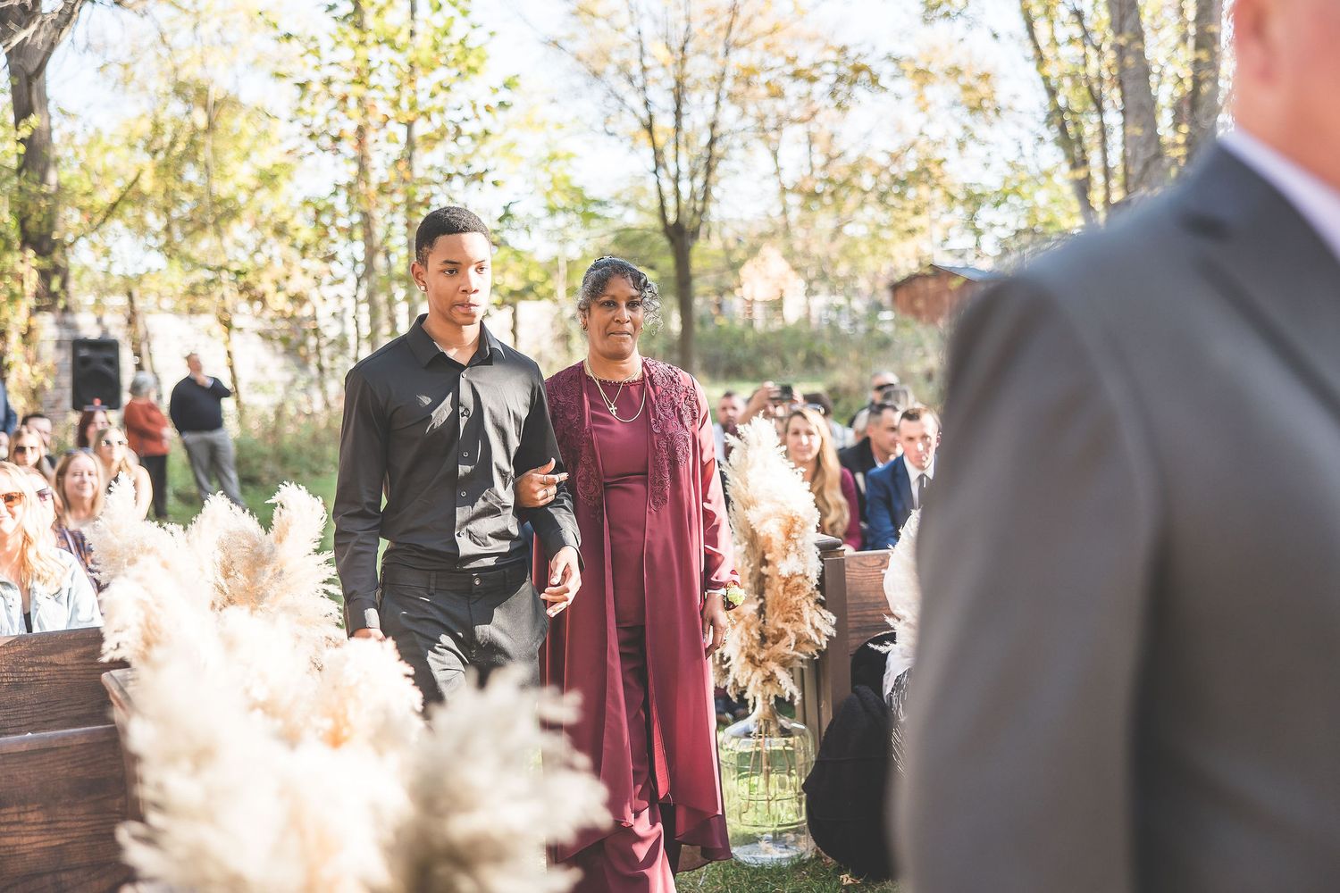 Wedding guests gather at an outdoor autumn ceremony with pampas grass decorations and fall foliage.